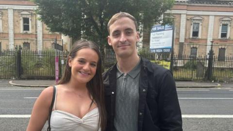 A brown haired woman and a taller, light-brown haired man stand in front of the derelict Crumlin Road Courthouse