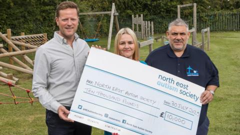 Robert Poole, Corrina McEwan and Tony Wedlake of the Teesside Family Foundation, standing on a grass field, with a wooden playground behind them, with a cheque which reads for a value of £10,000.