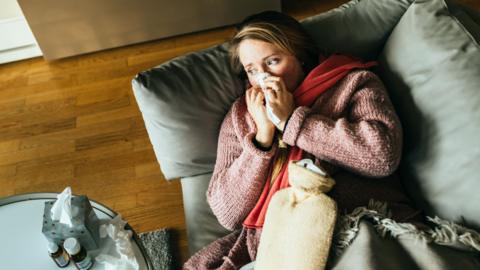 A woman lays on the sofa wrapped in blankets and holding a hot water bottle whilst blowing her nose.