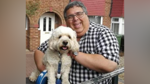 Councillor Richard Udall stands in a garden outside a red brick property. He wears glasses and a short-sleeved checked shirt. He is holding his white dog which looks like a poodle-cross.