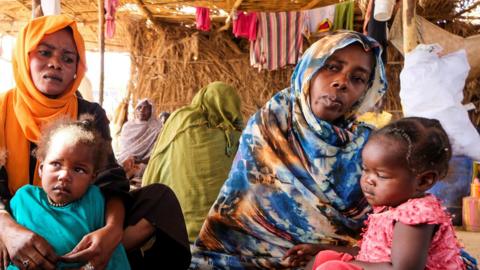 Two women wearing headscarves sit side by side, holding young children in their arms.