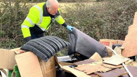 A council officer in a high-vis yellow jacket, wearing blue gloves and turning over household rubbish dumped beside a hedgerow