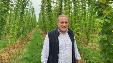 An image of Matthew King, the estate manager at the Hogs Back Brewery in Tongham, in Surrey. Matthew is standing in a field and is wearing a checked shirt and gilet. His hair is tied back. 