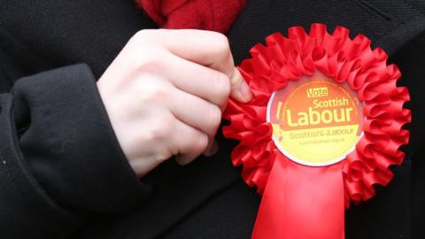 A person holds a red Labour rosette pinned to their black jacket 