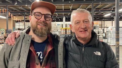 A man in an orange baseball cap and horned-rimmed glasses with his arm around a smaller man who is wearing a black jacket