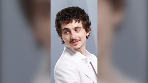 Actor Timothée Chalamet is wearing a white shirt and blazer, and looks back over his shoulder.