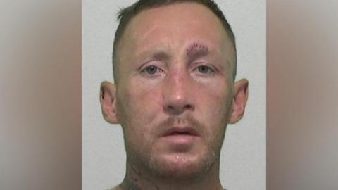 Mugshot of Roberts. He has faint ginger stubble and very short dark hair, a glazed expression and a large tattoo on his neck. He also has stitches above his left eyebrow.