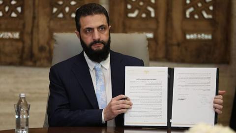 Syrian President Ahmed Sharaa in a navy suit, pale blue tie and white shirt holding up a physical copy of the agreement 