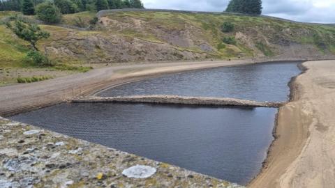 A thin stone bridge is spanning a body of water between two dry banks. The bridge rises slightly in the middle and wooden posts have been put in the banks at either side. In the distance there are hills and a forest to the far left.