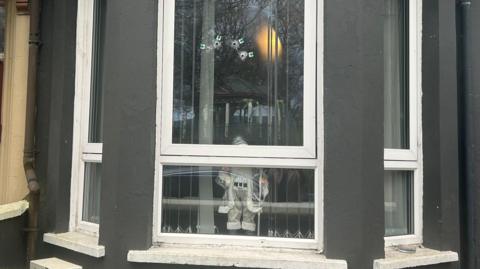 The window of a home. The walls are grey
There are four bullet holes in the window, beneath the bullet holes is a silver Santa decoration