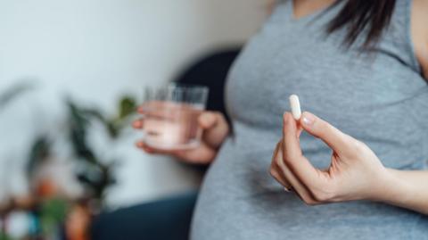 A pregnant women wearing a grey top is visible from the chest down, and holds a white pill in her left hand and a glass of water in her right (slightly blurred)