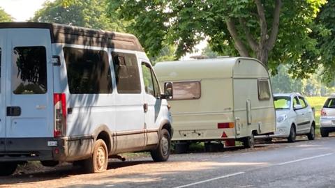 A row of vans parked along a road next to a grassy area in Bristol.