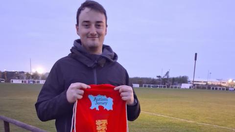 A picture of a man holding a red bag which reads 'Autism Jersey' on it. It is red and light blue in colour.