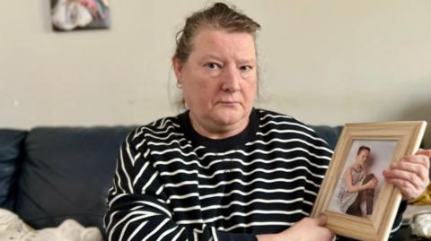 A woman sat on a sofa wearing a black and white stripy top while holding a photo in a frame of a girl