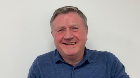 Dennis Jones smiling at the camera against a white wall, wearing a blue polo shirt.