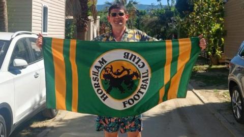 A man wearing sunglasses holding up a yellow and green towel with the Horsham FC badge on it.