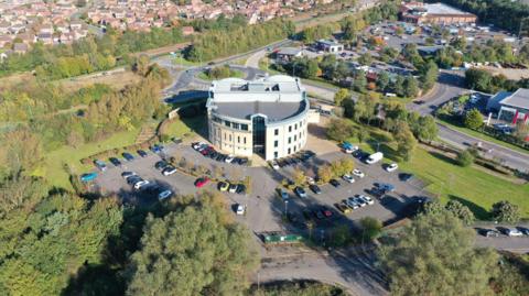 An aerial, drone-style photograph of a modern circular building surrounded by parking areas, greenery, and a mix of urban and suburban elements.
The environment combines developed infrastructure with large patches of woodland and open green space.
At the bottom of the image, dense clusters of mixed-height trees and shrubs fill the foreground.
A road runs through or near the lower part of the frame, partly obscured by the trees.