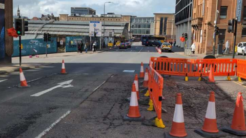 A road with part of it cordoned off by orange cones and barriers. Part of the road is slightly dipped and cracked