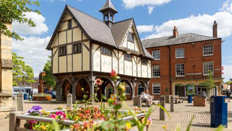 View of The Old Grammar School in Market Harborough, Leicestershire, with a bank to the right and flowers in the forefront on the image on a sunny day.
