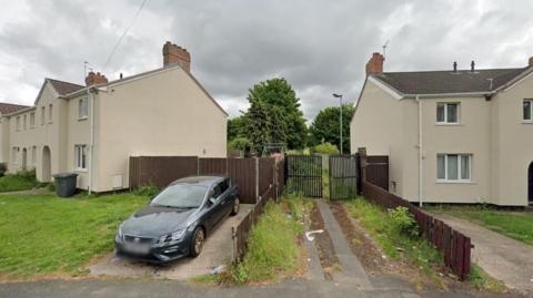 A pathway leads between two houses to a gate, partially open and which appears to lead to an area with greenery. A car is parked on the driveway of a nearby house.