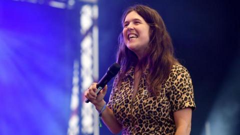 Rosie Jones in a leopard print jumpsuit looking off camera smiling and holding a microphone. She has brown hair and is standing on a stage.