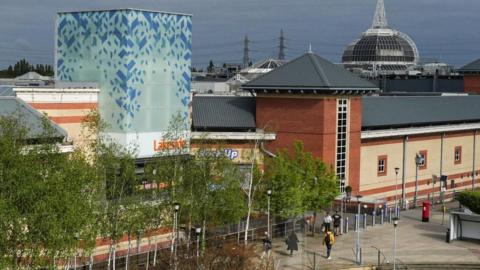 A general view of the large Lakeside Shopping Centre, which is a cream and orange brick building with a row of trees in front of it.