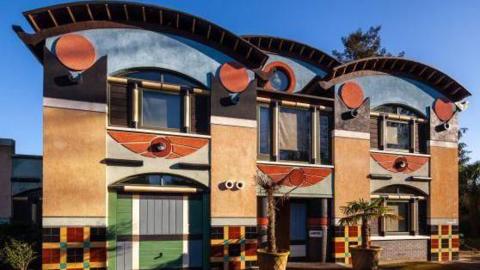 Exterior of a postmodern Egyptian-style property, featuring a winged solar disc over the main entrance, tartan pattern tiling along the base, and black columns topped with terracotta circles, representing the hieroglyph for the rising sun