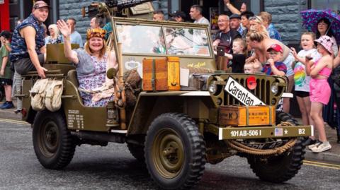 A lady in 1940s-style dress, apron and headgear waves as she sits in a mitliary jeep in a parade. Spectators line up on the pavement to watch.