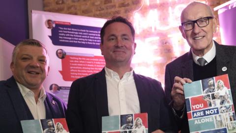 Nigel Seaman, Rob Parker and Terry Butcher stand together on a stage and smile at the camera. They each hold a copy of a book called You Are Not Alone and are wearing suit jackets with white shirts. 