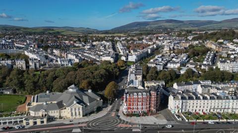 An aerial shot over Douglas, you can see a roundal in the foreground, on the left is the Villa Marina, there are lots of houses around and hills in the background.
