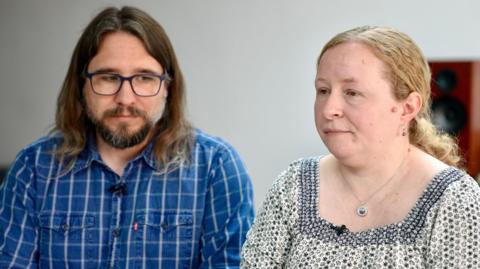 A close-up of Rob Miller and Katie Fowler seated side by side indoors. Miller is wearing glasses and a blue checked shirt and Fowler is wearing a patterned top and a pendant necklace. Both have clip-on microphones.