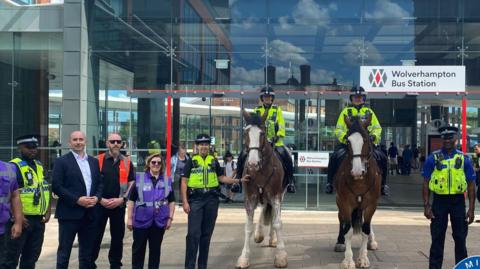 A group of West Midlands police and security colleagues wearing yellow hi-vis or purple t shirts are pictured next to two police horses outside Wolverhampton bus station