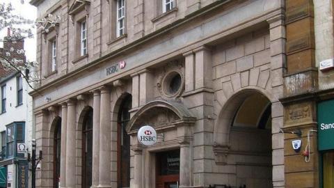 A HSBC bank in Kettering, showing the bank sign, the building is stone, and old, with stone columns and brick work. Buildings are either side.