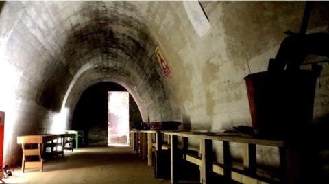This image shows a dimly lit, arched concrete air raid shelter, weathered walls and a single open doorway at the far end letting in light. Along one side, there are simple wooden benches and scattered objects.