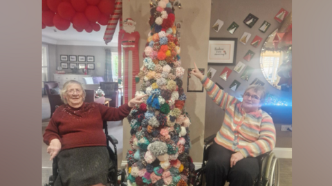 Vera Todd and Colette Roscamp are standing on each side of the colourful pom-pom Christmas tree with big star at its top.  They are pointing at pom-poms they have made. Vera is wearing a red cardigan and a grey skirt and has shoulder-length white hair. Colette is wearing black trousers and colourful striped cardigan. She has short grey hair. Both are wearing glasses. The rest of the room is decorated for Christmas with lights and red stars hanging from the ceiling. 