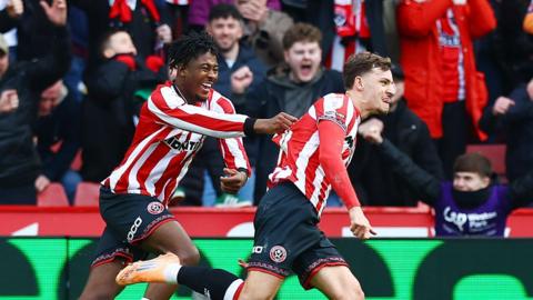 Harrison Burrows races away in celebration after scoring against Sheffield Wednesday as team-mate Andre Brooks grabs hold of the back of his shirt. In the background, fans in the stand at Bramall Lane shout with joy