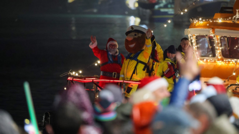 Father Christmas in his traditional red outfit stands next to a man wearing a lifeguard mascot costume, with yellow waterproofs and an oversized head with a brown beard on a lifeboat at night, while crowds of people wave from the shore.