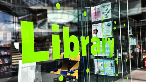 We can see a bright green 'Library' sign with letters stuck onto a glass panel. Behind it we can see the (out of focus) main Norwich library behind it.
