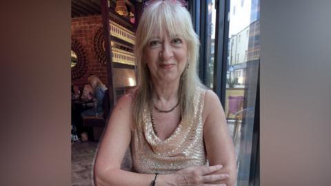 A woman with blonde hair and a fringe with pink-rimmed glassed on her head is smiling from someone across a table in what looks like a restaurant in the background. She is wearing a glittery beige and white top with a brown necklace.