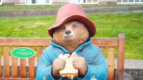 A statue of Paddington Bear sits on a brown wooden bench. It's wearing a red hat, blue coat and is holding a marmalade sandwich.