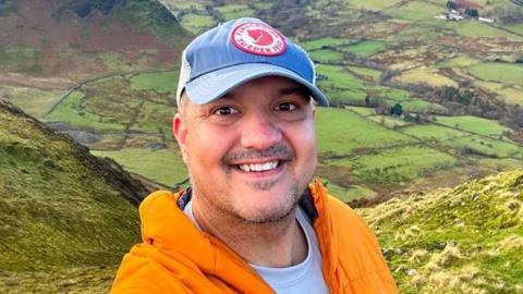 Jay Mistry stands a top of a fell in the Lake District. He stands in front of a grassy gorge. He is wearing an orange thermal fleece and a lilac t-shirt. He is wearing a light blue cap with a branded logo on it. 