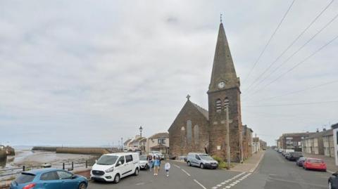 A general view of the road leading to Christ Church, with the harbour on the right-hand side. The church is at the bottom of the road and there are cars parked either side of the street. A family is walking past the church.