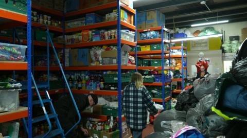 Volunteers organise stock amid rows of shelves stacked with groceries, and bin bags filled with sleeping bags and winter clothes