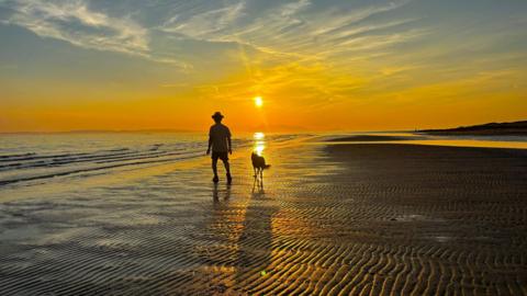 A silhouette of a man wearing a hat accompanied by a collie on a beach at sunset