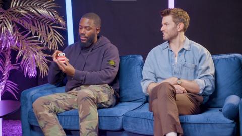 Mo Gilligan and Joel Dommett sit together on a blue sofa. Mo holds a model of the character teeth' and Joel smiles at it