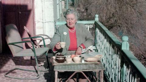 Agatha Christie, sitting at a table on an outdoor balcony, drinking tea