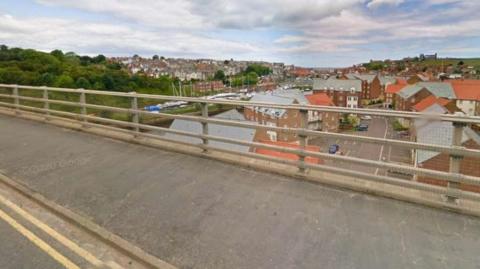 The foreground shows a tarmac footpath adjacent to a bridge rail of concrete uprights and a part of a road with double yellow lines marked on it. In the middle distance beneath the road, boats, trees and a river can be seen.