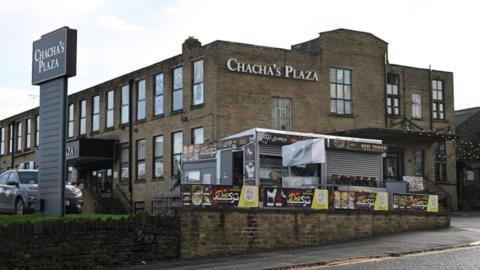 A view of a large two-storey brick building, seen from the other side of the road. There are lots of windows along one side of the building. Several signs can be seen on or around the building, saying "Chacha's Plaza". There is a street food van parked in front of the building.