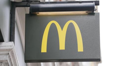 Signage for a branch of McDonald's. It is a dark grey sign with a yellow 'M' and it is it hanging on the wall of a white building.