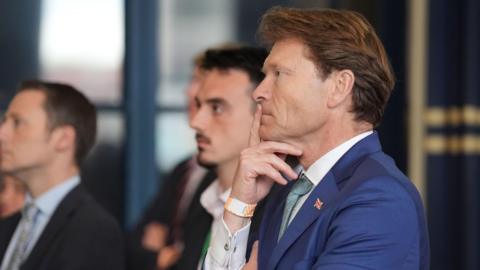 A side view of a man wearing a blue suit with a white shirt and green tie as results are announced at an election count. He has a finger to his mouth and is staring into the distance. There is a union flag badge on his suit lapel.